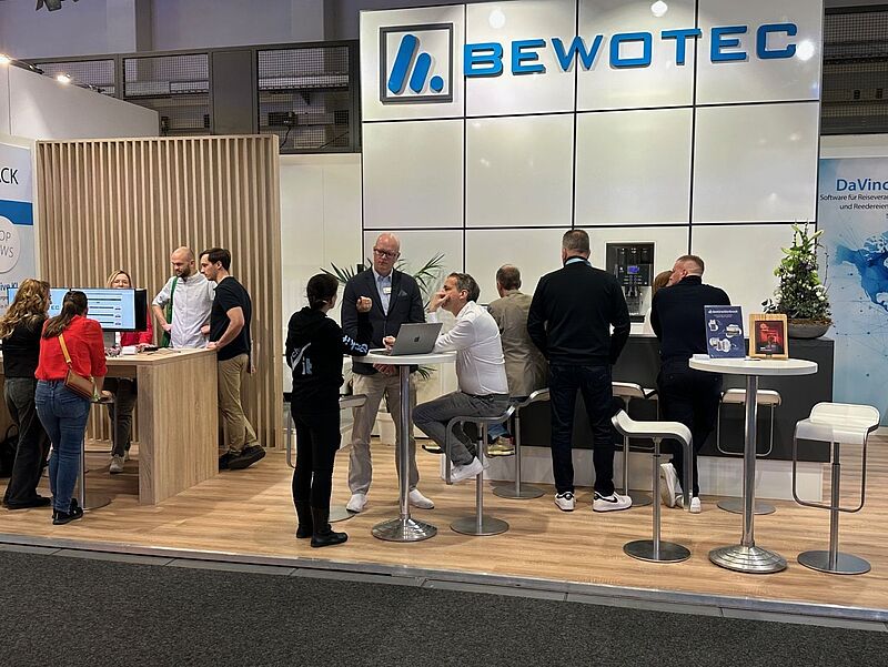 Mehrere Menschen stehen und sitzen an modernen Messeständen der Firma Bewotec auf einer Veranstaltung; es gibt Gespräche an Stehtischen und eine Theke mit Barhockern, der Firmenname „Bewotec“ ist groß auf der Wand hinter dem Stand sichtbar, einige Infomaterialien und ein Blumenarrangement sind auf den Tischen platziert.