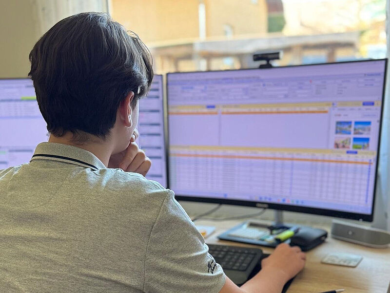 Ein junger Mann sitzt am Schreibtisch vor zwei Computerbildschirmen, auf denen Tabellen und Informationen angezeigt werden. Er trägt ein graues Polohemd und stützt sein Kinn nachdenklich auf eine Hand, während er mit der anderen Hand die Computermaus bedient. Im Hintergrund fällt Tageslicht durch das Fenster auf den Arbeitsplatz, auf dem Tastatur, Taschenrechner und weitere Büroutensilien liegen.
