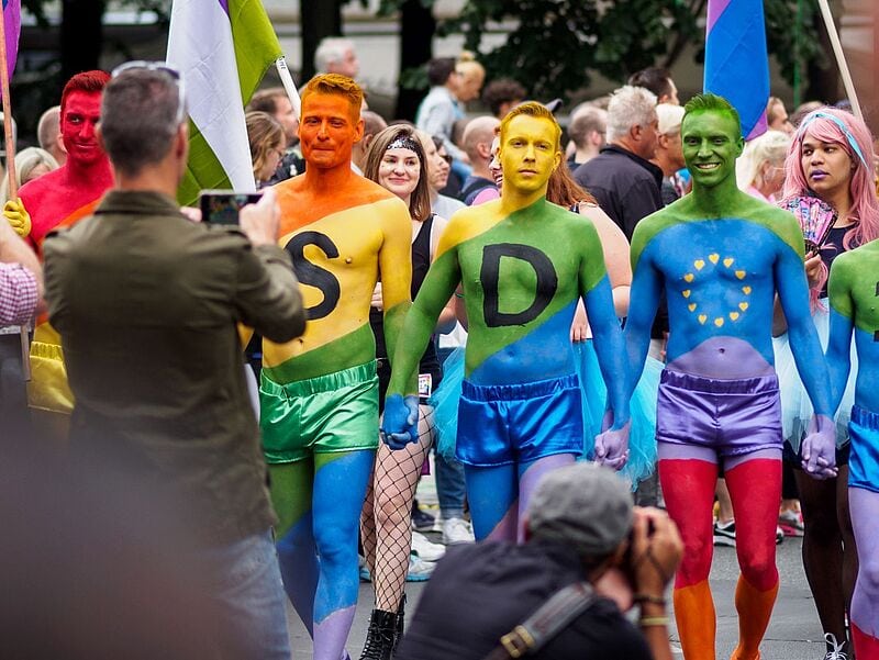 Eine Gruppe von Menschen nimmt an einer LGBTQ+-Parade teil. Im Vordergrund stehen fünf Männer mit bunt bemalten Oberkörpern und passenden kurzen Hosen und Tüllröcken in Regenbogenfarben. Zwei von ihnen haben große schwarze Buchstaben „S“ und „D“ auf die Brust gemalt, der dritte zeigt gelbe Sterne in Kreisform wie die EU-Flagge. Sie halten sich an den Händen und lächeln. Im Hintergrund fotografieren Zuschauer die Szene, während weitere Teilnehmende mit Regenbogen- und Pride-Flaggen zu sehen sind.