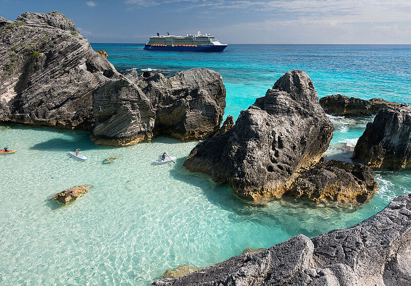 Auf Bermuda paddeln in seichtem türkisfarbenem Wasser und zwischen grauen Felsen zwei SUPler und blicken auf das offene Meer, wo im Hintergrund ein Kreuzfahrtschiff unterwegs ist. 