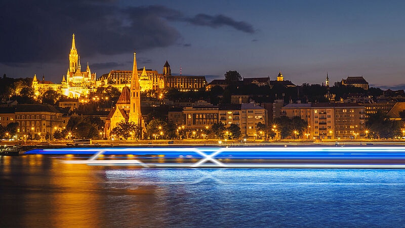 Ein stark stilisiertes, futuristisches Flusskreuzfahrtschiff aus leuchtendblauen Streifen und mit Markenzeichen X fährt im Abendlicht auf einem Fluss. Im Hintergrund sind erleuchtete Häuser und Kirchen zu sehen
