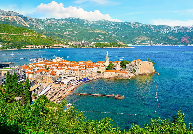 Mediterrane historische Stadt auf einer Landzunge umgeben von dunkelblauem Wasser, im Hintergrund ein grün bewachsenes Karstgebirge und blauem Himmel mit weißen Wolken. 