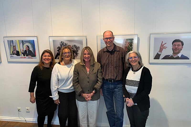 Gruppenfoto von fünf Personen vor weißer Galeriewand mit gerahmten Fotografien, professionell gekleidet in Business-Casual-Stil