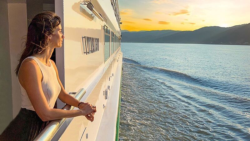 Eine Frau steht an einer Schiffsreling und schaut auf das Wasser. Man kann das Ufer erkennen