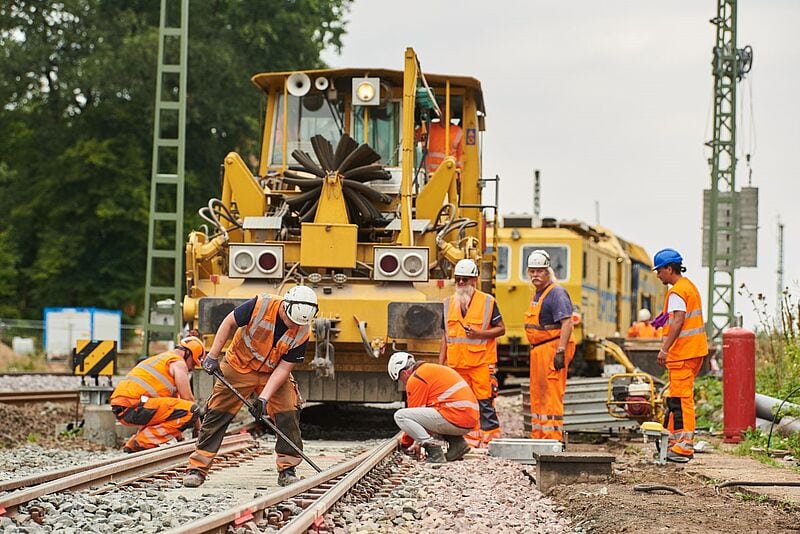 Wartungsarbeiten der Deutschen Bahn an Gleisanlagen: Ein Baustellen-Team in Sicherheitskleidung arbeitet mit schwerem Gerät an Schienen, eine gelbe Wartungsmaschine steht im Hintergrund