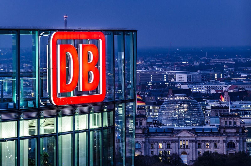 Das beleuchtete Logo der Deutschen Bahn an der Konzernzentrale in Berlin, im Hintergrund ist das Reichstagsgebäude zu sehen.