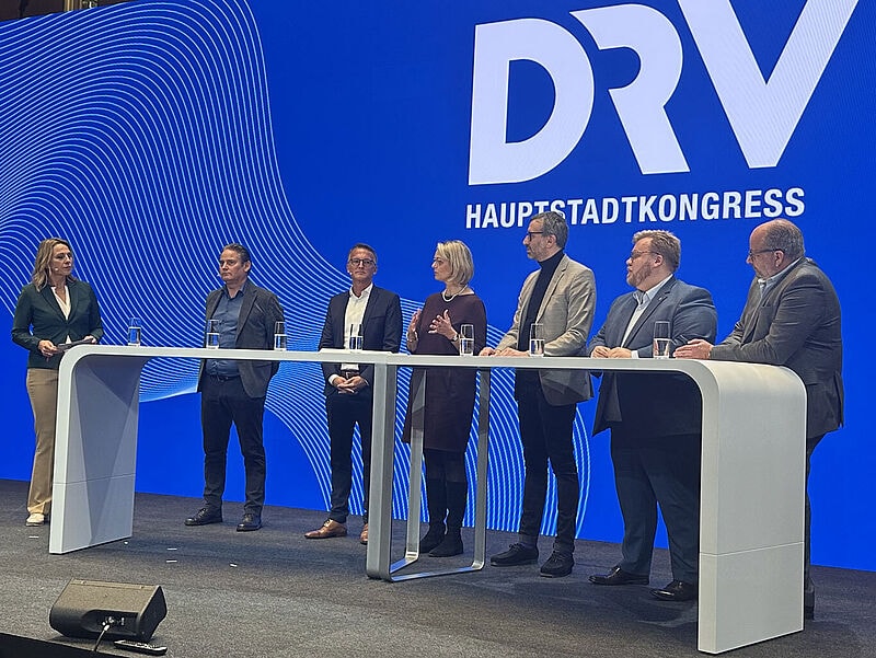 Podiumsdiskussion beim DRV Hauptstadtkongress: Sieben Teilnehmer stehen hinter weißem Pult vor blauem Hintergrund mit DRV-Logo.