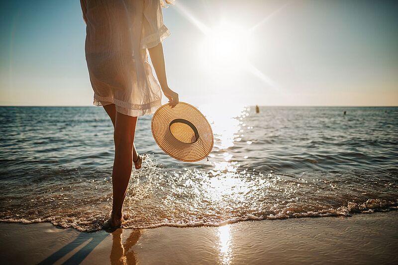 Frau in Rückansicht mit Sonnenhut in der Hand läuft am Meeressaum mit den Füßen im Wasser