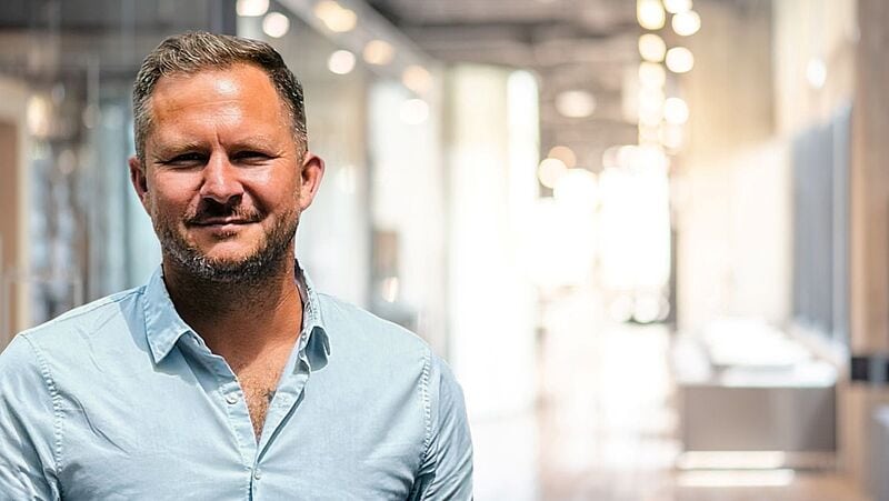 Mann mit kurzem Haar und Bart trägt ein hellblaues Hemd und steht in einem modernen, hell erleuchteten Büroflur