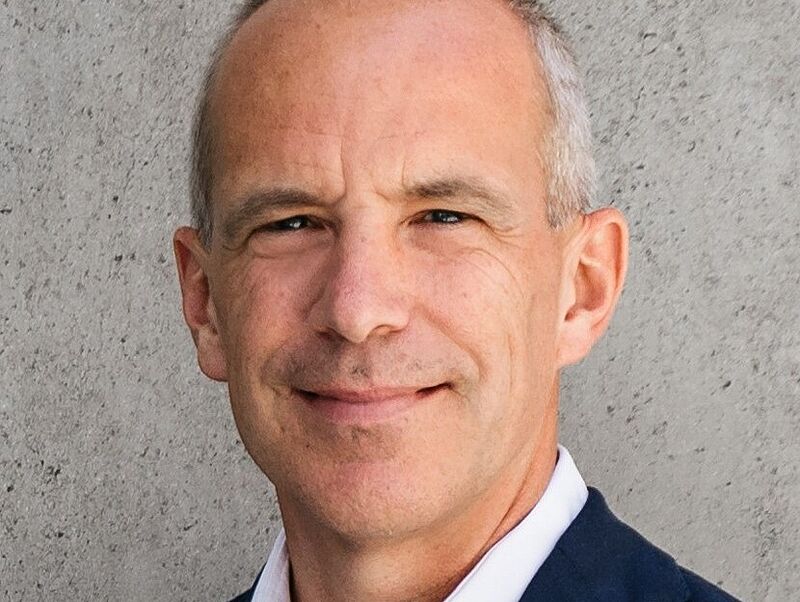 Porträt eines lächelnden Mannes in dunklem Sakko und weißem Hemd vor einer grauen Betonwand. Er hat kurze, graue Haare und wirkt professionell und freundlich.