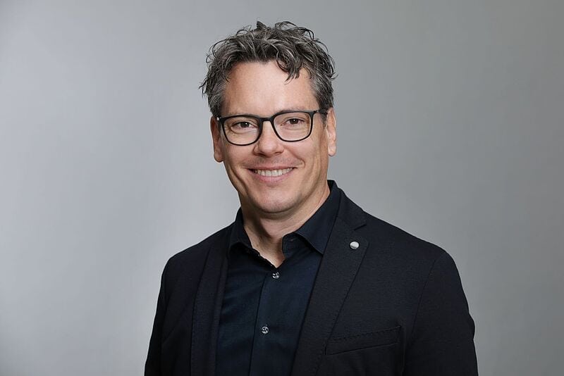 Ein Mann mit schwarzer Brille und schwarzem Hemd lächelt in die Kamera