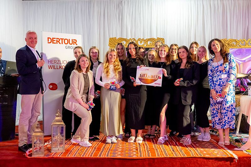 Eine Gruppe junger Menschen steht auf einem dekorierten roten Teppich vor einem weißen Vorhang bei einer Veranstaltung der Dertour Group. Im Vordergrund halten mehrere Teilnehmerinnen ein Plakat mit der Aufschrift „Campus Live – Tour 3“ sowie kleine Geschenke in den Händen. Links steht ein Mann mit Mikrofon vor einem Aufsteller mit dem Logo der Dertour Group und der Begrüßung „Herzlich Willkommen“. Die Atmosphäre ist festlich und freundlich.