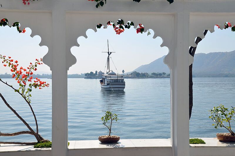 Blick durch kunstvoll verzierte weiße Bögen auf einen ruhigen See mit einem traditionellen Boot im Zentrum. Im Vordergrund stehen Blumentöpfe mit kleinen Pflanzen, und orangefarbene Blüten schmücken den oberen Rand des Rahmens. Im Hintergrund sind eine grüne Uferlinie und sanfte Hügel sichtbar.