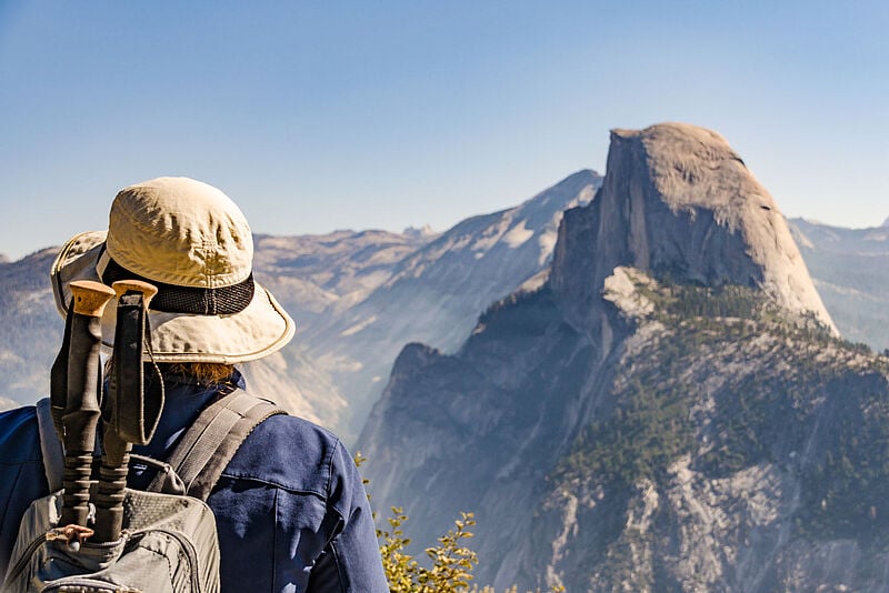Eine Wandertouristin mit Rucksack und Wanderstöcken steht im Yosemite-Nationalpark und betrachtet den Half Dome, der im Hintergrund unscharf zu sehen ist.