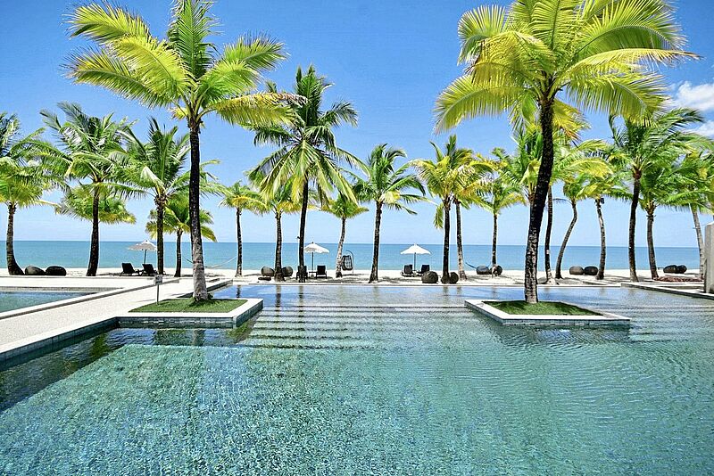 Tropisches Strandresort: Infinity-Pool umgeben von Palmen, im Hintergrund türkisblaues Meer und weißer Sandstrand.