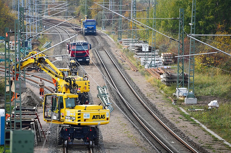 Baustelle zur Sanierung einer Bahnstrecke mit gelben Baggern im Vordergrund, roten und blauen Lastwagen auf einer Baustraße und Gleisanlagen mit Oberleitungsmasten.
