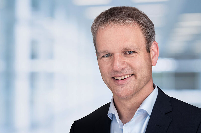 Ein Manager mittleren Alters mit weißem Hemd und dunklem Jackett lächelt in die Kamera vor unscharfem Hintergrund eines Bürogebäudes.