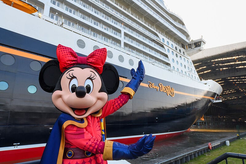 Eine mannshohe Minnie Maus in Superheldinnen-Kostüm steht vor dem neuen Kreuzfahrtschiff Disney Destiny und zeigt auf den dunklen Bug des Neubaus, der aus der Werfthalle gezogen und erstmals der Öffentlichkeit präsentiert wurde.