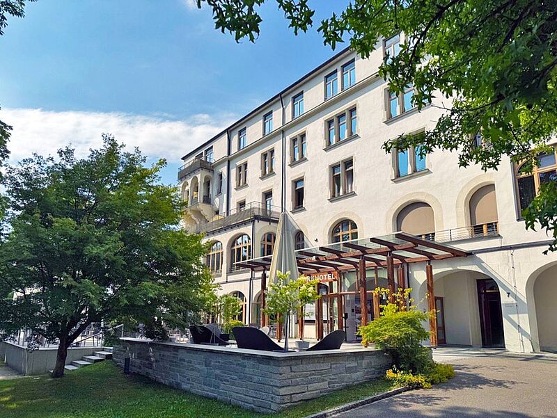 Außenansicht des historischen Dorint Hotels Jordanbad in Biberach an der Riß, Deutschland, umgeben von grünen Bäumen und blühenden Pflanzen unter blauem Himmel, ideal für Wellness- und Erholungsurlaub.