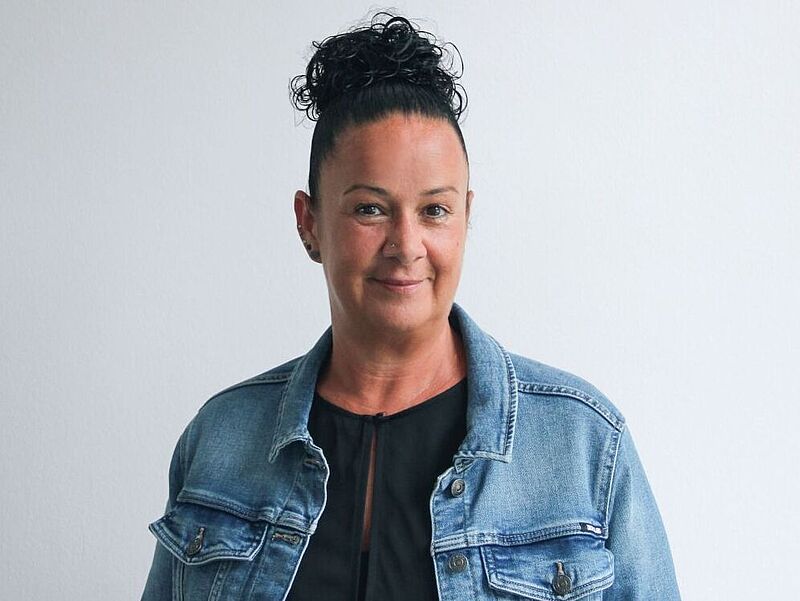 Frau mit dunklem, hochgestecktem lockigem Haar, trägt eine Jeansjacke über einem schwarzen Oberteil, steht vor einem hellen Hintergrund und lächelt freundlich in die Kamera.