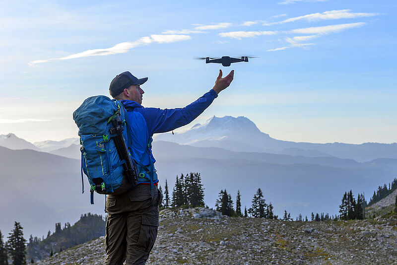 Wanderer mit blauem Rucksack steuert Drohne vor bergiger Landschaft mit schneebedeckten Gipfeln und Nadelwald.