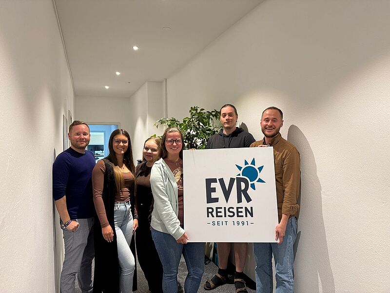 Sechs Personen stehen nebeneinander in einem hellen Büroflur. Zwei von ihnen halten ein Schild mit dem Logo „EVR Reisen – Seit 1991“. Alle lächeln in die Kamera.
