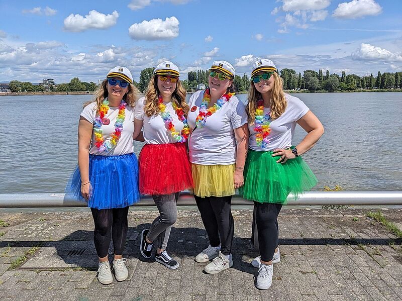 Vier Frauen stehen fröhlich nebeneinander am Ufer eines Flusses bei sonnigem Wetter. Sie tragen weiße T-Shirts, bunte Tüllröcke in Blau, Rot, Gelb und Grün sowie Matrosenmützen und bunte Hawaiiketten. Jede von ihnen trägt zudem Sonnenbrillen und bequeme Schuhe. Im Hintergrund ist das Wasser, eine Baumreihe und ein leicht bewölkter Himmel zu sehen. 