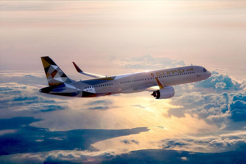 Ein Flugzeug von Etihad Airways des Typs Airbus A321 LR fliegt bei Sonnenuntergang über Wolken. Am Leitwerk befindet sich ein geometrisches Muster, am Rumpf der Schriftzug von Etihad. Die Lichtstimmung am Himmel ist dramatisch.