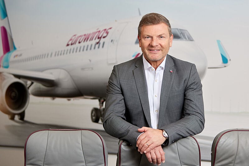 Ein Manager mittleren Alters mit grauem Jackett und weißem Hemd steht über einen Flugsitz gelehnt vor einem unscharfen Hintergrund, der ein Flugzeug der Airline Eurowings zeigt