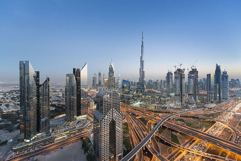 Luftbild der Skyline von Dubai am Abend mit beleuchteten Hochhäusern und einem verzweigten Netz autobahnähnlicher Straßen mit viel Verkehr.