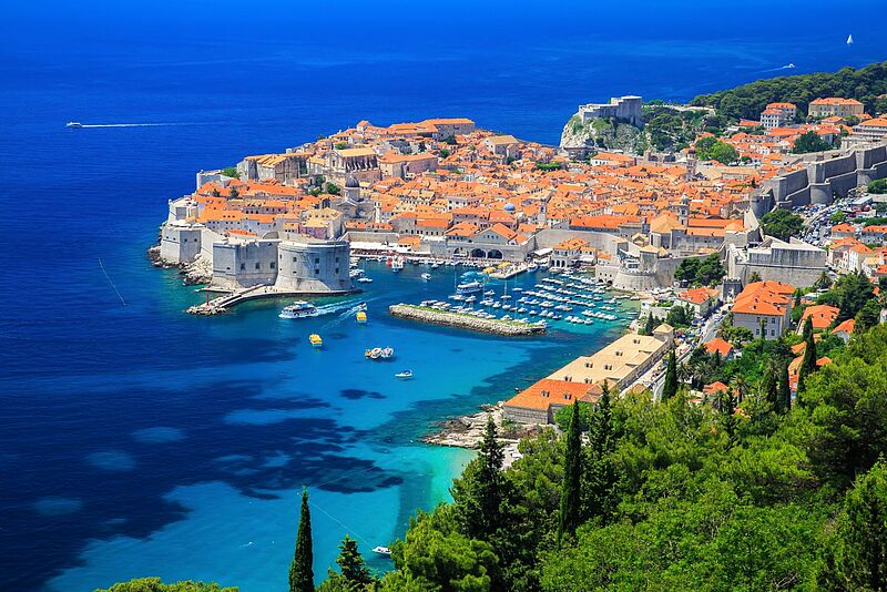 Panoramablick auf die Altstadt von Dubrovnik in Kroatien, umgeben von einer massiven mittelalterlichen Stadtmauer. Die roten Ziegeldächer der historischen Gebäude heben sich deutlich vom tiefblauen Wasser der Adria ab. Im Hafen liegen zahlreiche Boote und Yachten, während die grüne Vegetation im Vordergrund einen schönen Kontrast zur urbanen Struktur bietet. Am Horizont erstreckt sich das offene Meer unter klarem, wolkenlosem Himmel.