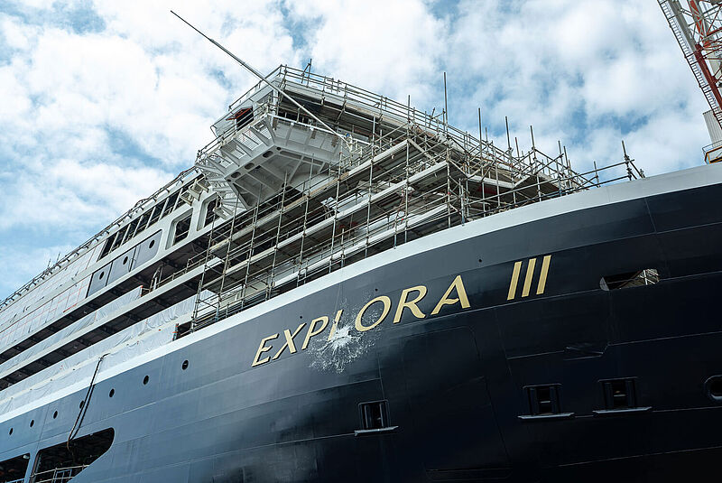 Am dunkelblauen Bug eines neugebauten Kreuzfahrtschiffs zerschellt eine Taufflasche direkt am Schiffsnamen Explora III, der in goldfarbenen Lettern an der Schiffswand prangt.