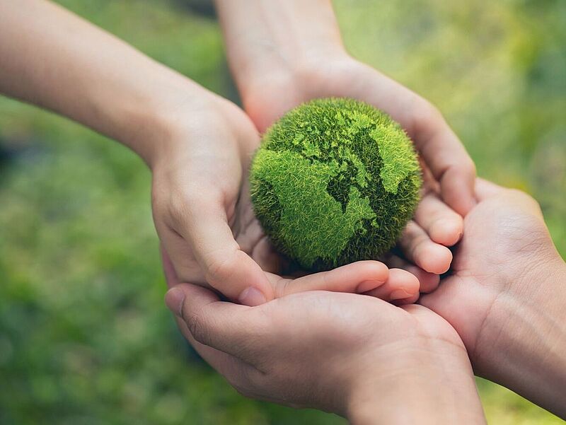 Mehrere Hände halten gemeinsam eine grüne, moosbedeckte Kugel in Form der Erde – ein Symbol für Umweltschutz, Nachhaltigkeit und gemeinschaftliche Verantwortung für unseren Planeten.“