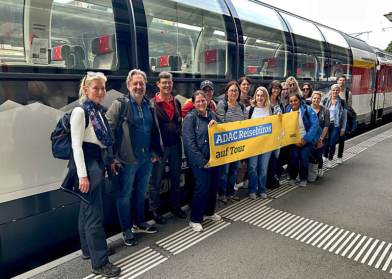 Eine Reisegruppe von etwa 15 Personen steht auf einem Bahnsteig vor einem Zug mit großen Panoramafenstern. Sie halten vor sich ein Transparent mit der Aufschrift „ADAC Reisebüros auf Tour“.