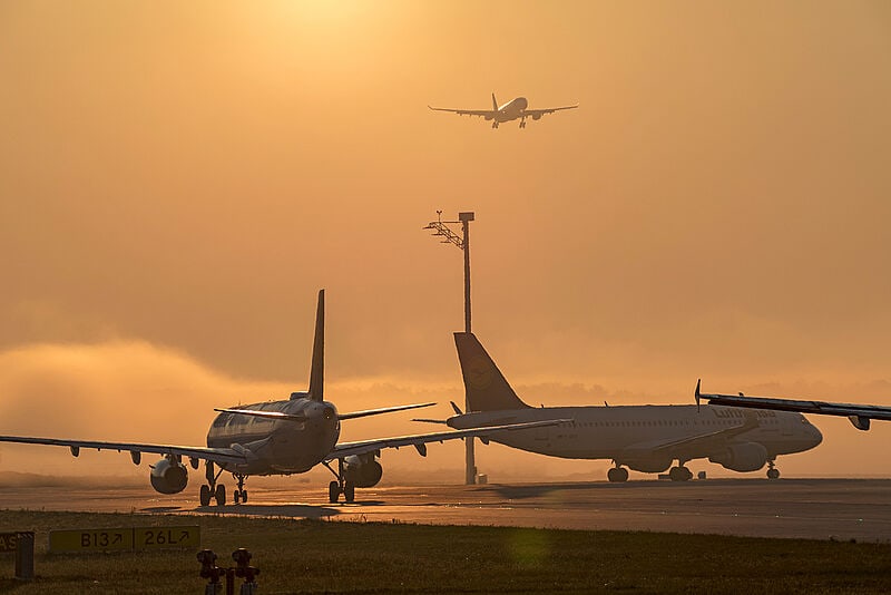 Goldene Stunde mit orangefarbenem Himmel auf den Start- und Landebahnen des Flughafens München. Während zwei Flugzeuge am Boden bereitstehen, nähert sich ein weiteres im Landeanflug.
