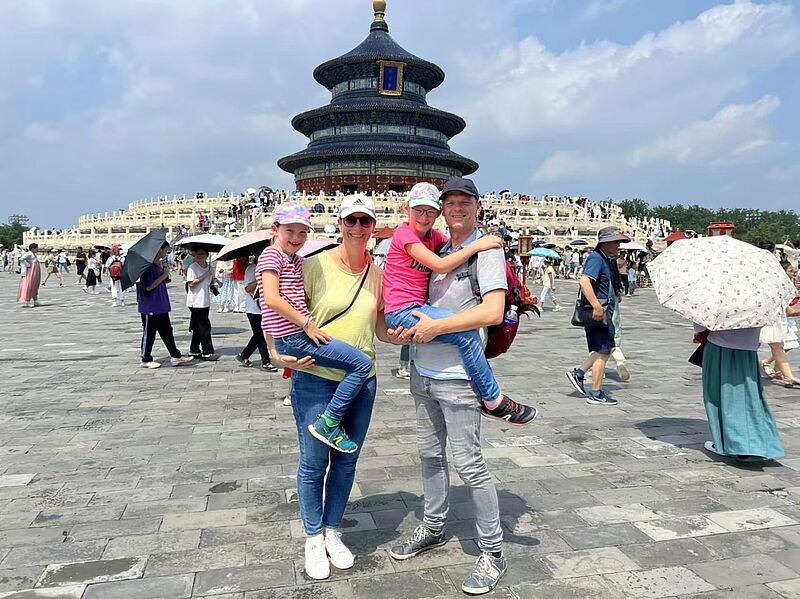 Eine Familie mit zwei Kindern posiert fröhlich vor dem Himmelstempel (Tempel des Himmels) in Peking, China. Die Eltern halten jeweils eines der Kinder auf dem Arm. Im Hintergrund sind zahlreiche Besucher sowie das markante, runde Gebäude des Himmelstempels mit seinem blauen, dreistufigen Dach zu sehen. Es ist ein sonniger Tag mit leicht bewölktem Himmel, viele Menschen schützen sich mit Schirmen vor der Sonne.