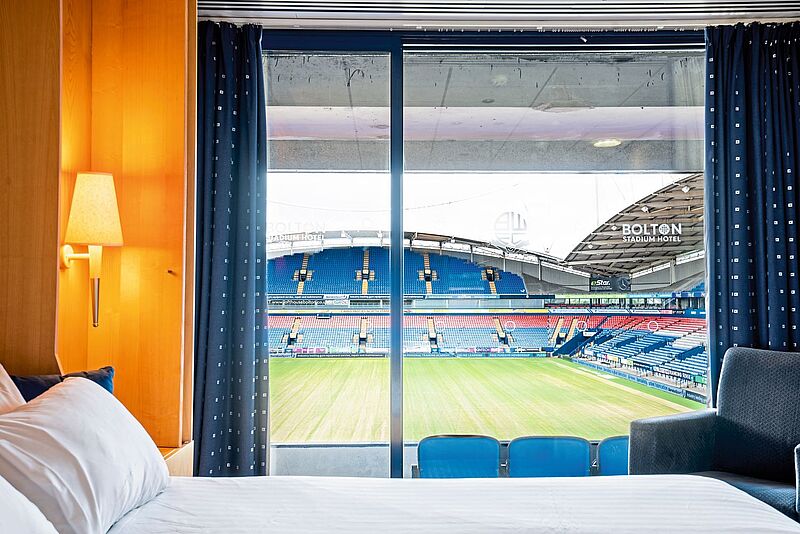 Blick in ein Hotelzimmer, hinter einem mit weißer Bettwäsche bezogenen Bett, über dem eine Nachttischlampe angeknipst ist, sieht man durch ein Fenster in den Innenraum eines Stadions