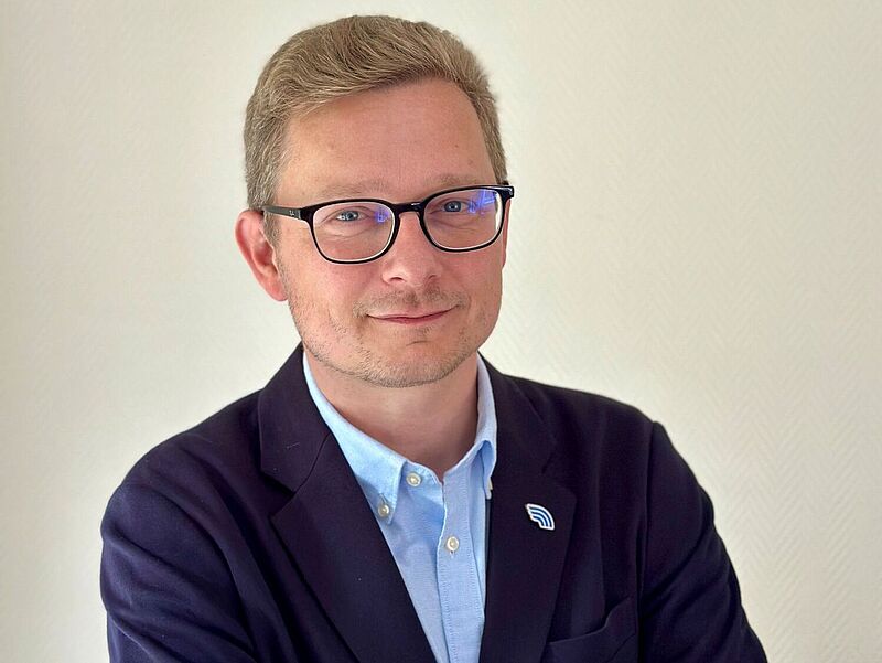 Ein Mann mit blonden, kurzen Haaren und Brille steht vor einer hellen Wand. Er trägt ein dunkelblaues Sakko mit einem blauen Hemd und hat die Arme verschränkt. Am Revers trägt er eine Anstecknadel mit einem stilisierten Logo.