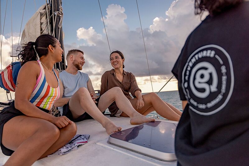 Drei junge Erwachsene sitzen entspannt auf dem Deck eines Segelbootes und unterhalten sich lachend, während im Hintergrund die Sonne über dem Meer untergeht. Eine vierte Person im Vordergrund trägt ein T-Shirt mit dem Logo von G Adventures.