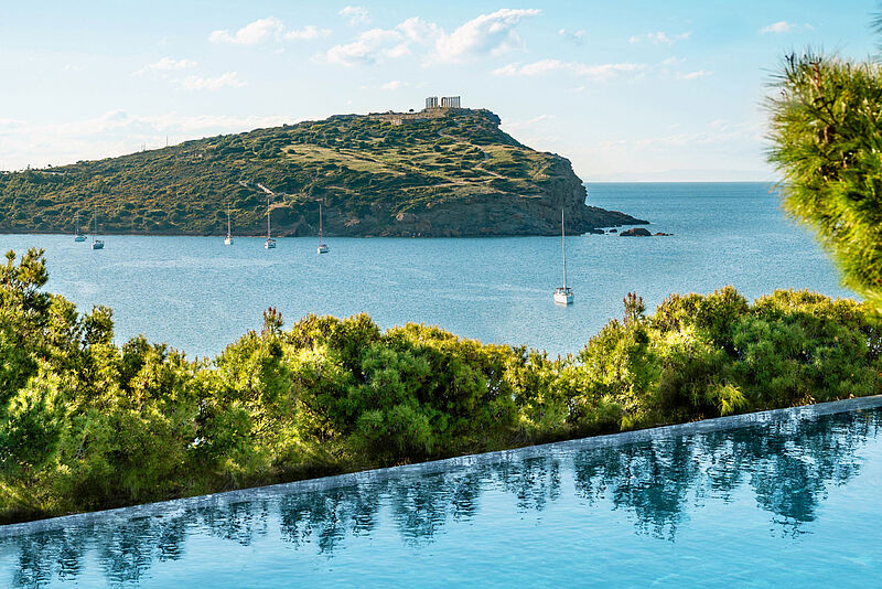 Infinity-Pool mit Blick auf das Meer, mehrere Segelboote und eine grüne Halbinsel mit dem antiken Tempel des Poseidon auf der Spitze, unter blauem Himmel.