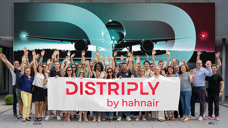 Das Team der neuen Produktmarke Distriply von Hahnair steht lächelnd und mit erhobenen Armen vor einem großen und stilisierten Flugzeug-Wandbild. Die Mitarbeiter halten vor sich ein Banner mit dem Firmenlogo.