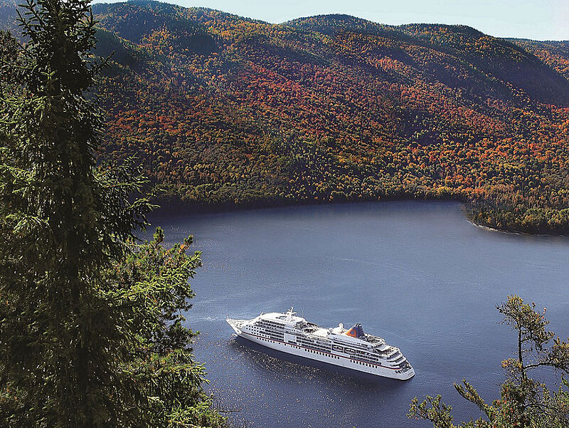 Ein weißes Kreuzfahrtschiff liegt zur Zeit des Indian Summer in einer von farbenprächtigen Wäldern umgebenen Bucht. Die Sonne scheint.
