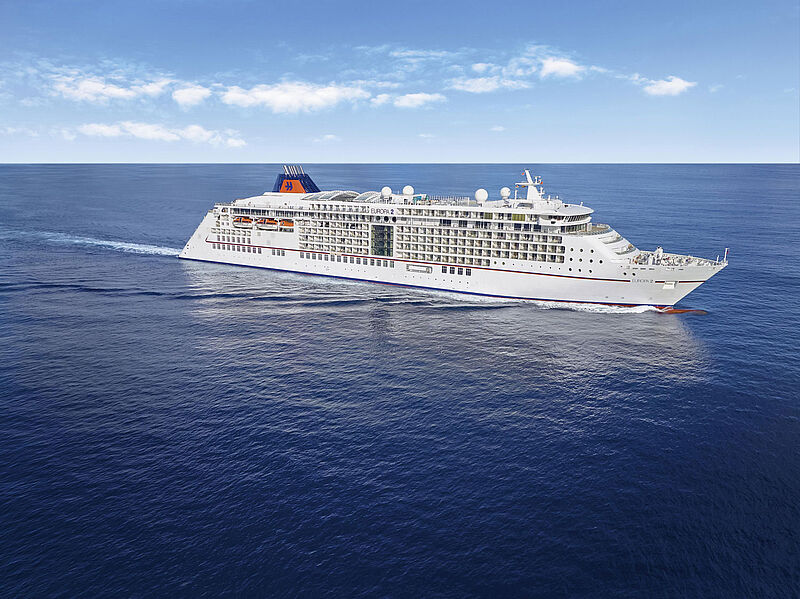 Das kleine Kreuzfahrtschiff Europa 2 mit weißem Rumpf und blau-orangenem Querstreifen an der Schiffswand fährt auf offenem blauem Meer. Die Sonne scheint.
