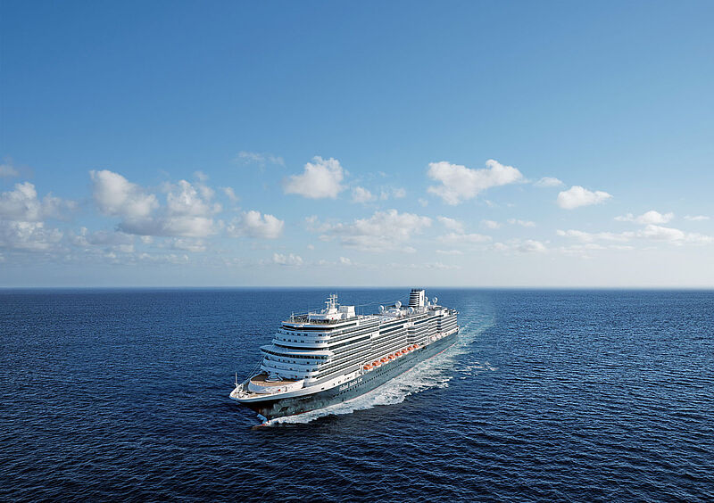 Frontale Luftaufnahme eines Kreuzfahrtschiffes auf offener See. Der dunkelblaue Rumpf hebt sich nur leicht vom Meer ab, darüber sind hellblauer Himmel und weiße Wolken zu sehen.