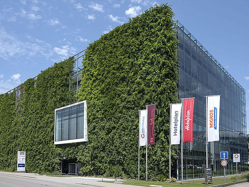 Modernes Bürogebäude mit vollständig begrünter Fassade auf der linken Seite und gläserner Front auf der rechten Seite. Vor dem Gebäude stehen vier Fahnenmasten mit den Logos von Hotelplan, Migros und weiteren Unternehmen. Der Himmel ist leicht bewölkt.