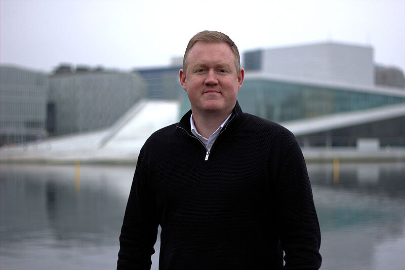 Porträtfoto von einem Mann mittleren Alters mit kurzen braunen Haaren, der in schwarzem Cardigan an einem Ufer steht. Im Hintergrund ist ein futuristisch anmutendes, graues Gebäude zu erkennen.
