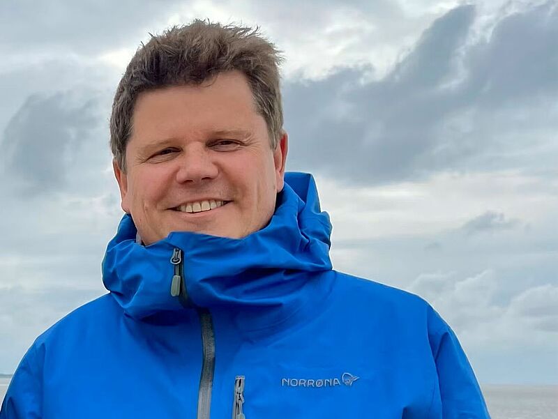 Ein Mann mittleren Alters mit braunem Haar steht in einen blauen Parka gehüllt vor weißen Wolken.