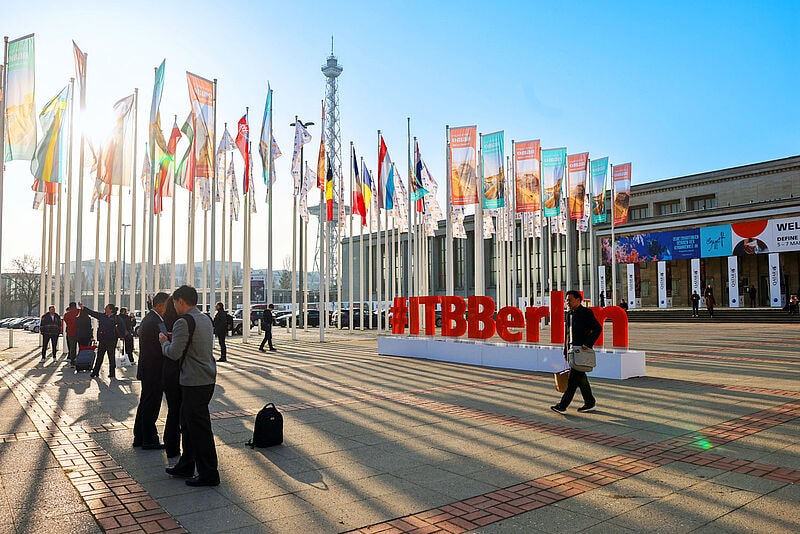 Eingangsbereich ITB Berlin mit Flaggenallee, rotem ITB-Logo und Messegebäude im Morgenlicht, Besucher im Vordergrund