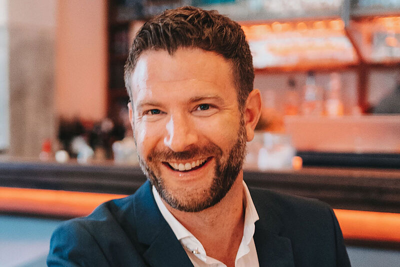 Professionelles Porträt in Restaurant-Setting: Person in dunkelblauem Blazer und weißem Hemd lächelt herzlich vor warmem Ambiente.