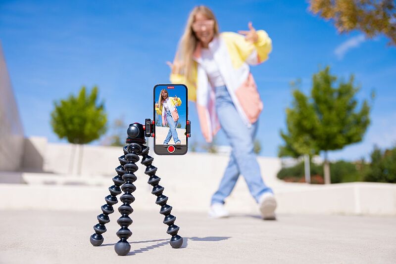 in Smartphone auf einem flexiblen Stativ filmt eine junge Frau, die draußen vor blauem Himmel posiert. Die Frau trägt eine bunte Jacke und Jeans und ist unscharf im Hintergrund zu sehen, während ihr Bild scharf auf dem Handydisplay erscheint.